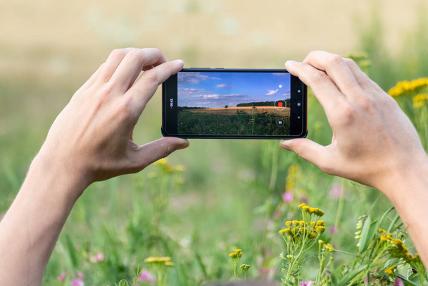 両手でスマートフォンを水平に持ち、夏の麦畑の写真を撮る夏の日当たりの良い自然景観 - 写真・画像