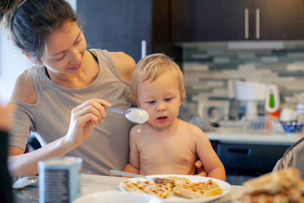 Mère souriante regardant le petit fils et le nourrissant après avoir cuit des biscuits debout à table dans la cuisine. Bonne maman attentionnée et adorable enfant manger le dîner. - Photo, image