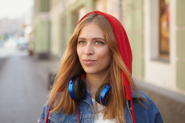 Beautiful caucasian young woman about 25 years old in hoodie and with earphones is walking during the street in Saint-Petersburg and posing for photos. White young lady in the city center. - Foto, immagini