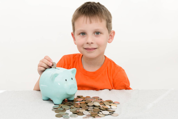 A preschool boy in an orange T-shirt sits and puts coins in a piggy bank. Concept - the child learns to save, save for a dream, and recounts savings. The horizontal photo looks into a frame, smiles - Photo, Image
