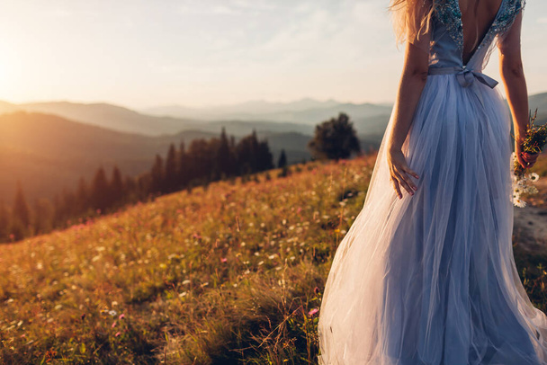 Schöne Braut im blauen Hochzeitskleid in den Bergen bei Sonnenuntergang. Die junge Frau genießt die sommerliche Landschaft der Karpaten mit einem Blumenstrauß. Rückseite - Foto, Bild