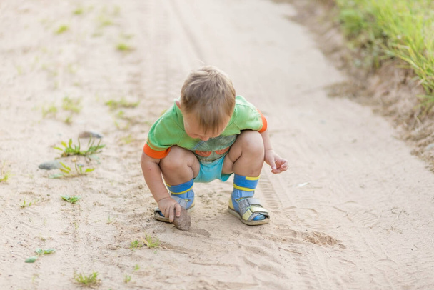 Boy draws with a stone on the sand while squatting on a sandy village road. - 写真・画像