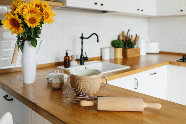 Dishes and utensils on kitchen table, ready to cook. white simple modern kitchen in scandinavian style, kitchen details, wooden table, sunflowers bouquet in vase on the table - Foto, afbeelding