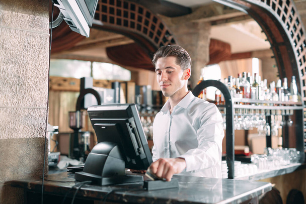 small business, people and service concept - happy man or waiter in apron at counter with cashbox working at bar or coffee shop. - Fotografie, Obrázek