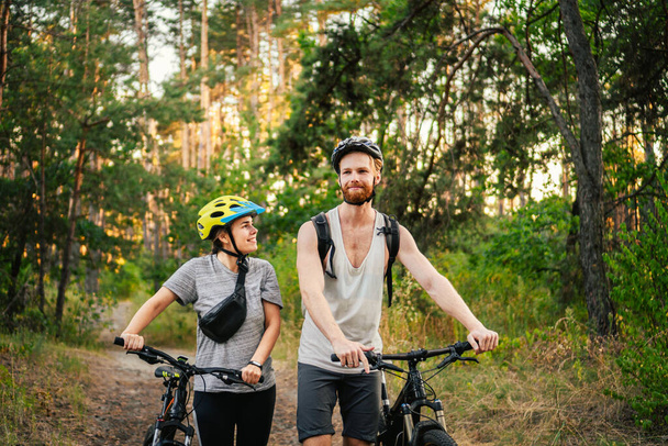 Una joven pareja de ciclistas caucásicos caminan y empujan sus bicicletas de montaña a lo largo de la carretera forestal en el parque. Fin de semana deportivo activo. Deportiva gente hermosa con una bicicleta en el campo. - Foto, imagen