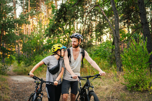 Vrolijk echtpaar duwen fietsen en wandelen langs de bosweg. Gelukkig stel met fiets lopend door park en pratend en lachend. Kaukasische jonge man en vrouw fietsen in het bos. - Foto, afbeelding