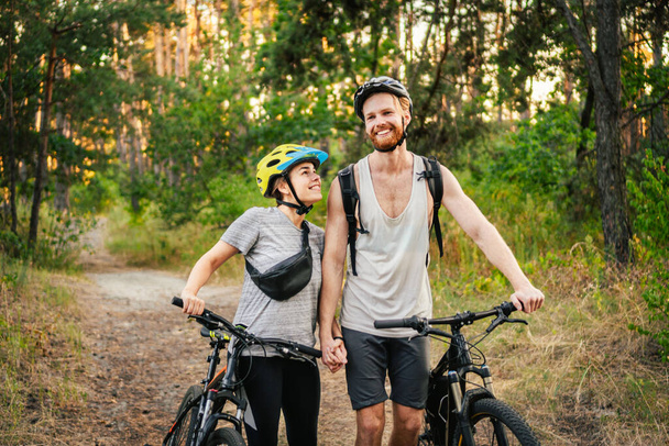 Ein munteres Pärchen schiebt Fahrräder und läuft die Forststraße entlang. Glückliches Paar mit Fahrrad, das durch den Park läuft und redet und lacht. Kaukasischer junger Mann und Frau auf Fahrrädern im Wald. - Foto, Bild