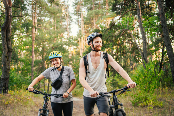 Nuoret valkoihoiset pyöräilijät kävelevät ja työntävät maastopyöränsä puistossa olevaa metsätietä pitkin. Aktiivinen urheiluviikonloppu. Urheilullinen kauniita ihmisiä pyörä maaseudulla. - Valokuva, kuva