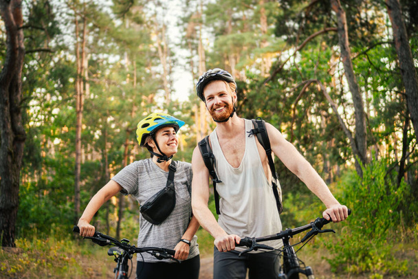 Χαρούμενο ζευγάρι που σπρώχνει ποδήλατα και περπατάει στο δασικό δρόμο. Ευτυχισμένο ζευγάρι με ποδήλατο να περπατάει στο πάρκο και να μιλάει και να γελάει. Καυκάσιος νεαρός άνδρας και γυναίκα ιππεύουν ποδήλατα στο δάσος. - Φωτογραφία, εικόνα
