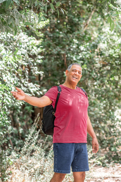 Portret van een gelukkige activiteit atleet senior man met een rugzak, genieten van een prachtig uitzicht. Gezonde oude man op een wandeltocht. Mannelijke gids in het bos doet een hand uitnodigend gebaar. - Foto, afbeelding