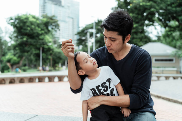 A man in black t-shirt embrace his son while he sleep at daytime in the park. - Photo, Image