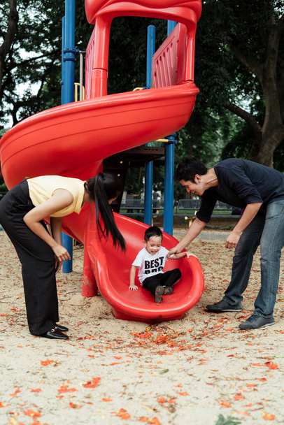 Apa és anya áll a végén a piros csúszó a játszótéren a parkban, hogy elkapják a fiukat. - Fotó, kép