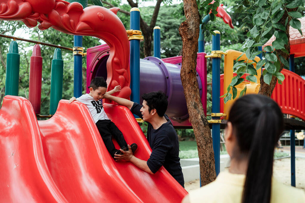 ママを見てパパと息子は公園の遊び場で赤いスライダーで遊ぶ. - 写真・画像