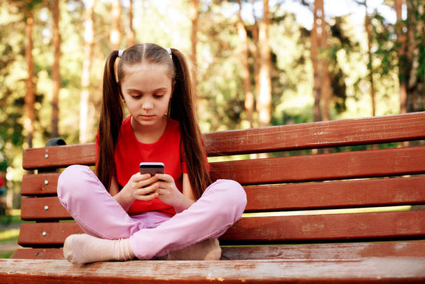 Little Beautiful School Girl Sitting On A Park Bench Texting On The Smart Phone. - Photo, Image