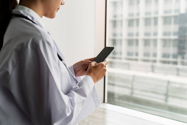 Image recadrée du smartphone entre les mains d'une femme médecin alors qu'elle se tenait à la fenêtre et utilisait son téléphone. - Photo, image