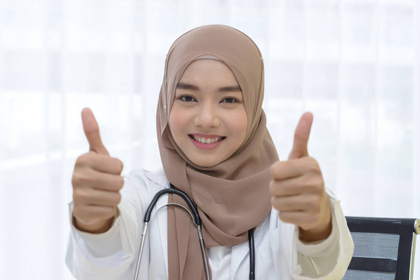 Κοντινό πορτρέτο της χαμογελαστή νεαρή μουσουλμάνα γυναίκα ιατρός με στηθοσκόπιο δείχνει αντίχειρες επάνω υπογράψει στην κλινική - Φωτογραφία, εικόνα