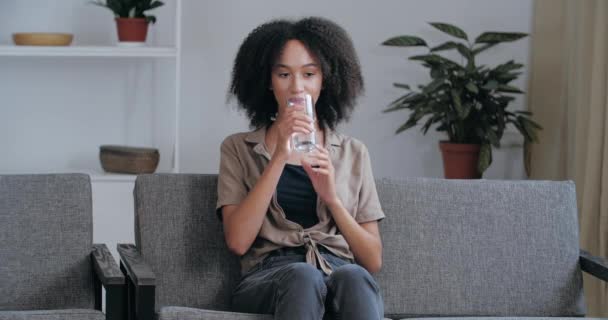 Young African American woman sitting on couch at home in stuffy room resting relaxing, quenching her thirst with cold clean still mineral water on hot summer day, feeling refreshing relief health care - Footage, Video