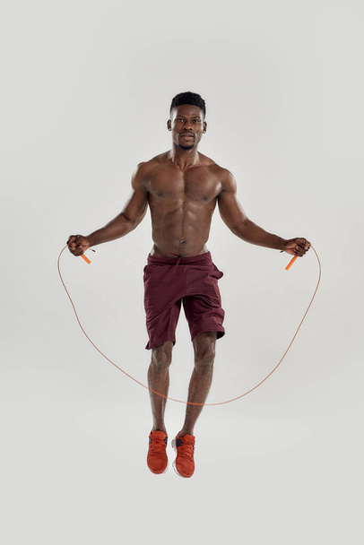 Full length shot of young muscular african american man looking at camera, while doing exercises with jumping rope isolated over grey background. Sports, workout, bodybuilding concept - Valokuva, kuva