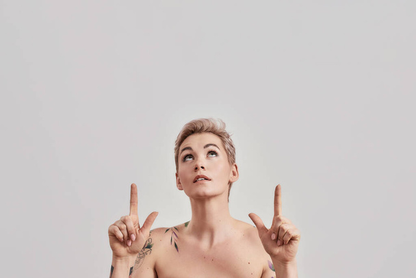 Look here. Portrait of half naked tattooed woman with short hair looking up pointing index fingers at the upper part isolated over light background - Photo, Image