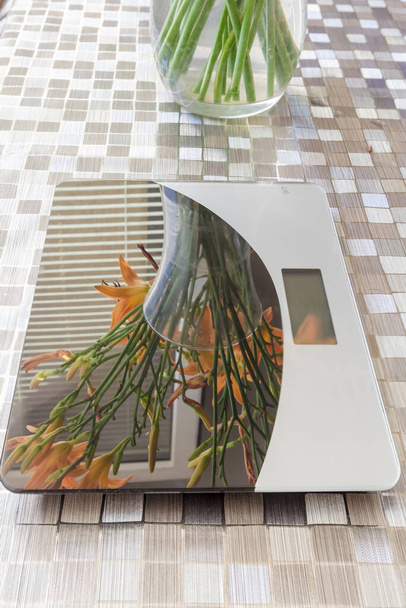 Пустая бытовая цифровая кухонная шкала с букетом цветов, отражающих на рабочей поверхности, на столе, покрытом салфеткой - Фото, изображение