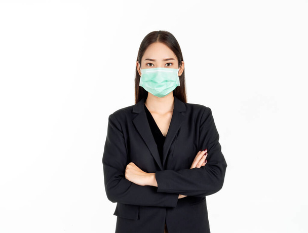Una bella donna d'affari asiatica che indossa una maschera medica verde e un vestito nero, in piedi con le braccia incrociate e guardando la fotocamera isolata su sfondo bianco. - Foto, immagini
