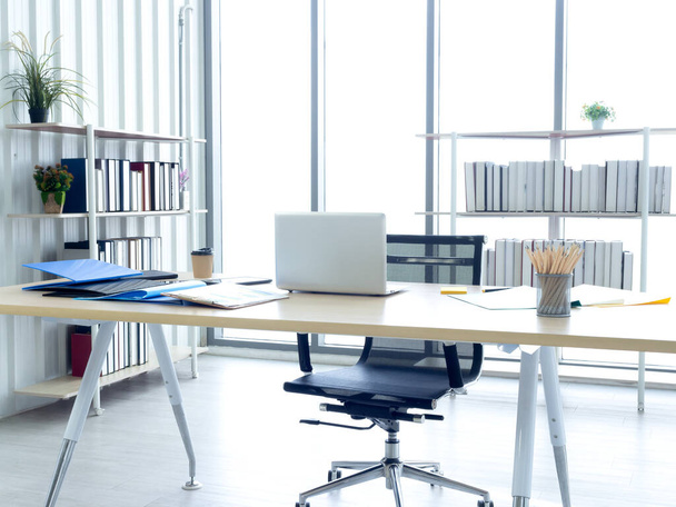 Modern office interior with huge windows. Well-lit workplace with laptop computer, pencils, coffee cup and document file folders on desk with empty chair and bookshelf in the back. - 写真・画像