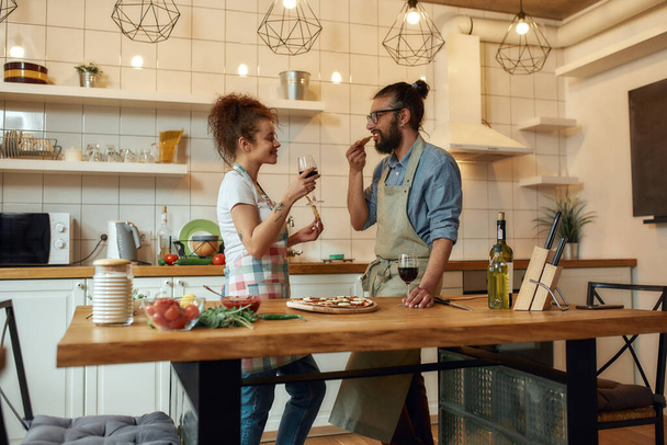 Enjoy the moment. Young man and woman in apron eating freshly baked pizza and drinking wine while standing in the kitchen. Love, relationships concept - Fotografie, Obrázek