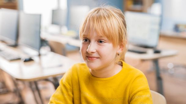 Portret słodkiej dziewczynki z blond włosami siedzącej przy biurku, uśmiechnięta szczęśliwie. Mądra dziewczynka z uroczym uśmiechem Siedząca w klasie. Zdjęcia portretowe z bliska - Zdjęcie, obraz