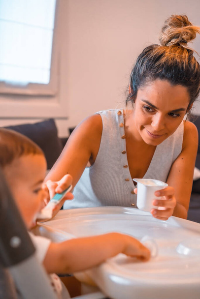 Young Caucasian mother feeding her son a yogurt while sitting in the highchair. Teleworking and caring for your child, a new normal after the coronavirus pandemic, covid-19. - Foto, Imagem