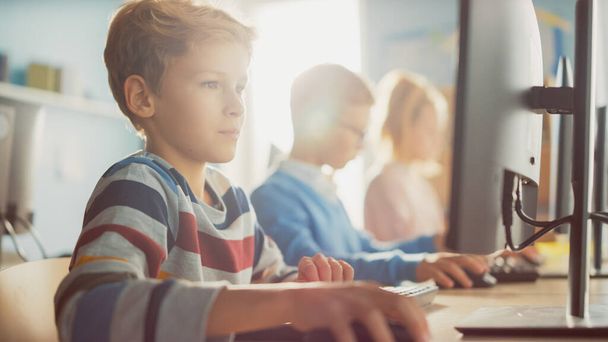 Elementary School Classroom: Boy Uses Personal Computer, Learning How to Use Internet Safely, Programming Language for Software Coding. Schoolchildren Getting Modern Education. Shot with warm Filter - 写真・画像
