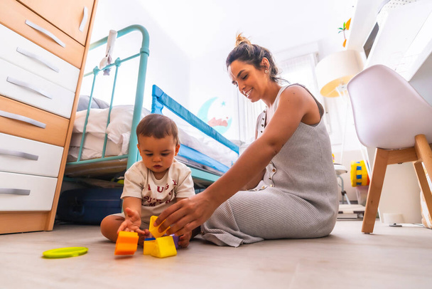 Young Caucasian mother playing with her in the room with toys. Baby less than a year learning the first lessons of her mother. Mother playing with her son playing sitting on the floor - Foto, Bild