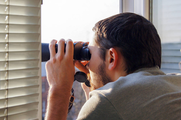 Junger Mann steht mit Fernglas durch ein Glasfenster und beobachtet etwas in der Ferne. - Foto, Bild