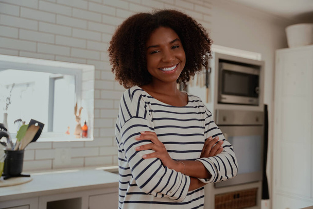 Portrait of cheerful young african woman standing in modern kitchen with folded arms looking at camera - Fotografie, Obrázek