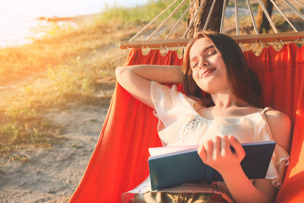 Dışarıda hamakta dinlenirken kitap okuyan genç bir kadın. - Fotoğraf, Görsel