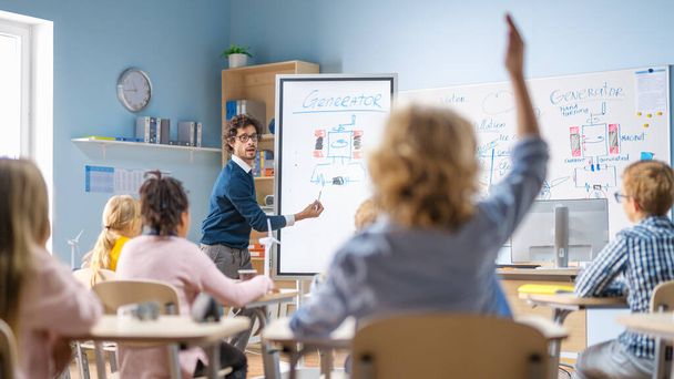 Brilliant Schoolboy Listens Attentively to His Teacher Explaning Lesson and Raises His Hand with a Question. In Elementary School with Group of Bright Multiethnic Kids Learning Science. Back View Shot - 写真・画像