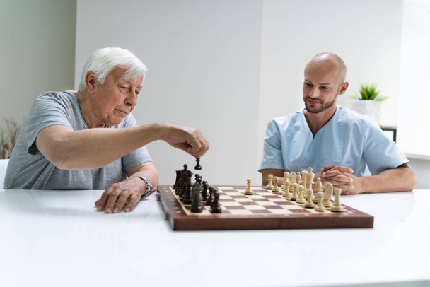 Elderly Senior Playing Chess With Caregiver At Home - Фото, изображение