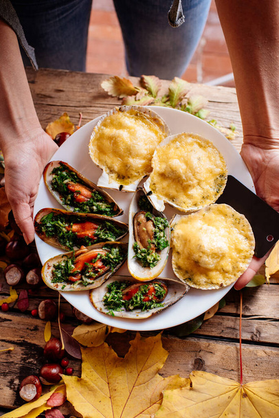 Baked mussels and scallops baked under cheese on white plate in hands of woman on wooden table with autumn leaves and chestnuts. - Foto, imagen