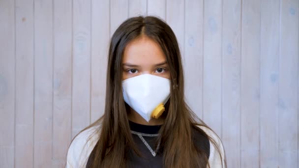 Egy tinédzser lány, aki orvosi védőmaszkot visel, a kamerába néz. Covid-19, koronavírus járvány. Portré gyönyörű barna tini lány orvosi védő maszk felett fény szürke fal háttér - Felvétel, videó
