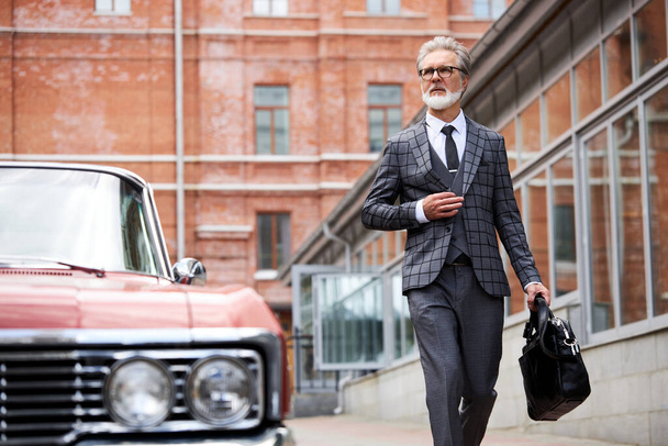 灰色の髪で髭を生やした上品な老人が屋外でバッグを持って歩く - 写真・画像