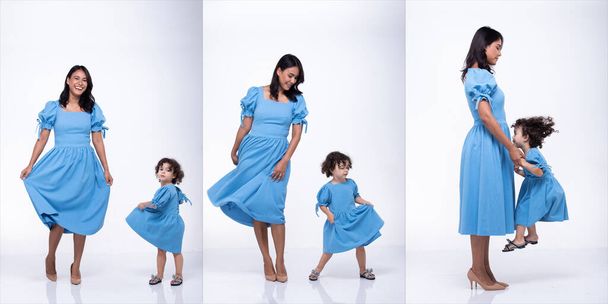 Ασιάτισσα μητέρα και καυκάσια κόρη στέκονται και φορούν το ίδιο μπλε φόρεμα μπλούζας μαζί. Το κοριτσάκι κρατάει το χέρι της μαμάς και χαμογελάει με αγάπη. Απομονωμένο λευκό φόντο - Φωτογραφία, εικόνα