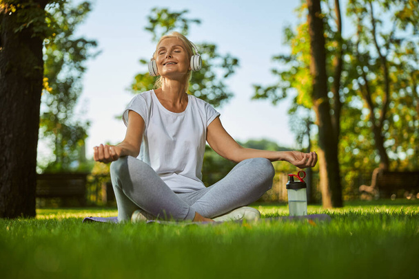 Charming woman in headphones practicing yoga in park - Фото, изображение