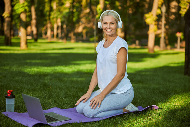 Cheerful woman in headphones using laptop in park - Fotó, kép