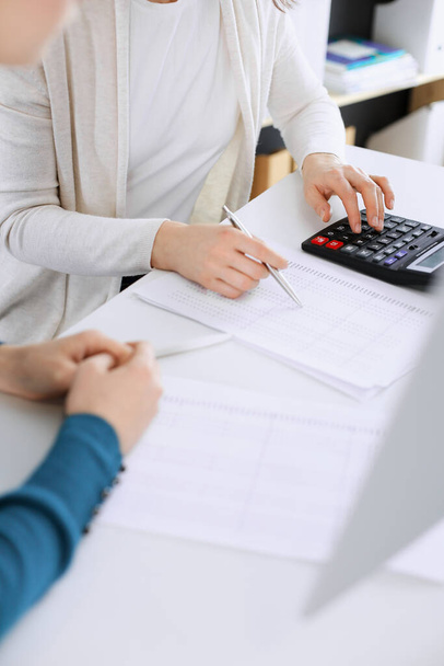 Accountant checking financial statement or counting by calculator income for tax form, hands closeup. Business woman sitting and working with colleague at the desk in office toned in blue. Tax and - 写真・画像