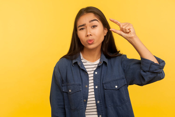 Too small! Portrait of displeased girl in denim shirt showing a little bit gesture, dissatisfied with size measuring low scale, minimum inch or centimeter. studio shot isolated on yellow background  - Foto, imagen