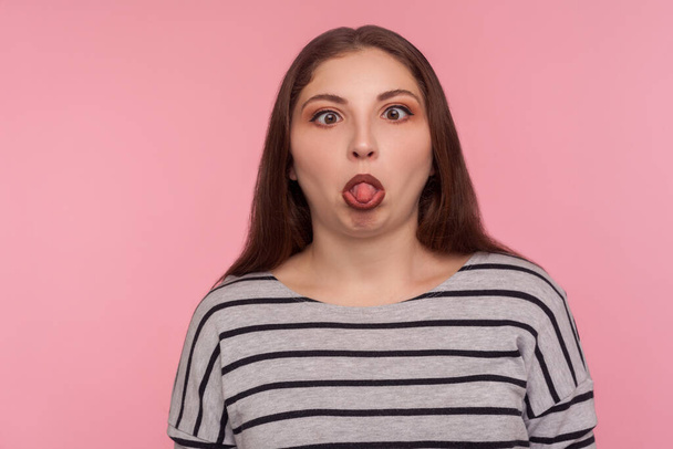 Portrait of funny dumb amusing woman in striped sweatshirt standing with crossed eyes, showing tongue out and making silly brainless facial expression. indoor studio shot isolated on pink background  - Fotó, kép