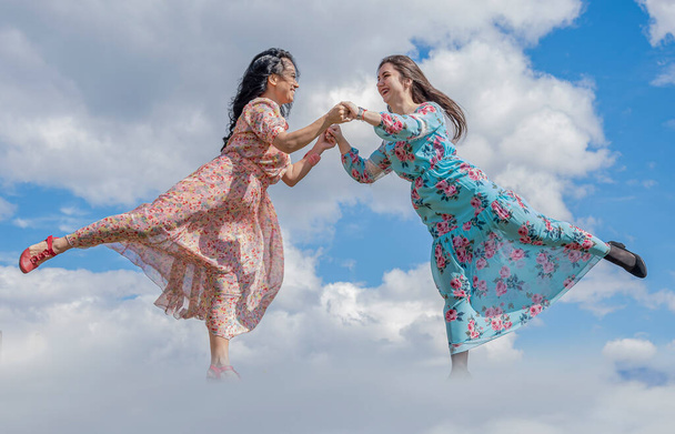 Dwie szczęśliwe dziewczyny w jasnych letnich sukienkach wzięły się za ręce, patrzą na siebie na tle błękitnego nieba. - Zdjęcie, obraz
