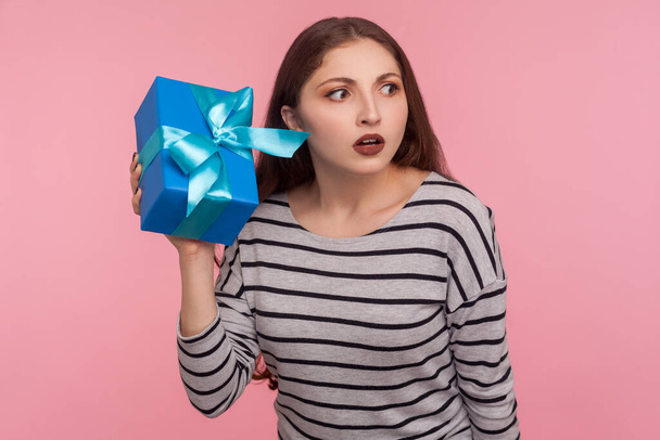 What's inside? Portrait of woman in striped sweatshirt holding gift box near ear and listening, guessing birthday surprise, in anticipation of interesting present. indoor studio shot, pink background  - Photo, image