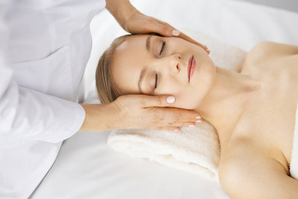 Beautiful caucasian woman enjoying facial massage with closed eyes in spa salon. Relaxing treatment in medicine and Beauty concept - Zdjęcie, obraz