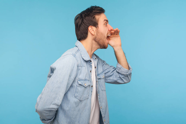 Attention, important message! Side view of aggressive man in worker denim shirt yelling loud into hands shaped like megaphone, proclaiming announcement. indoor studio shot isolated on blue background  - Foto, Imagem