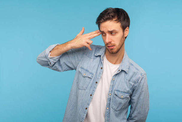 Suicide gesture. Portrait of frustrated desperate young man in worker denim shirt pointing finger gun to head, feeling depressed and shooting himself. indoor studio shot isolated on blue background  - Φωτογραφία, εικόνα
